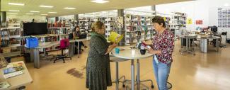 Intérieur d'une bibliothèque avec une bibliothécaire à son bureau et deux personnes qui lisent debout autour d'une table